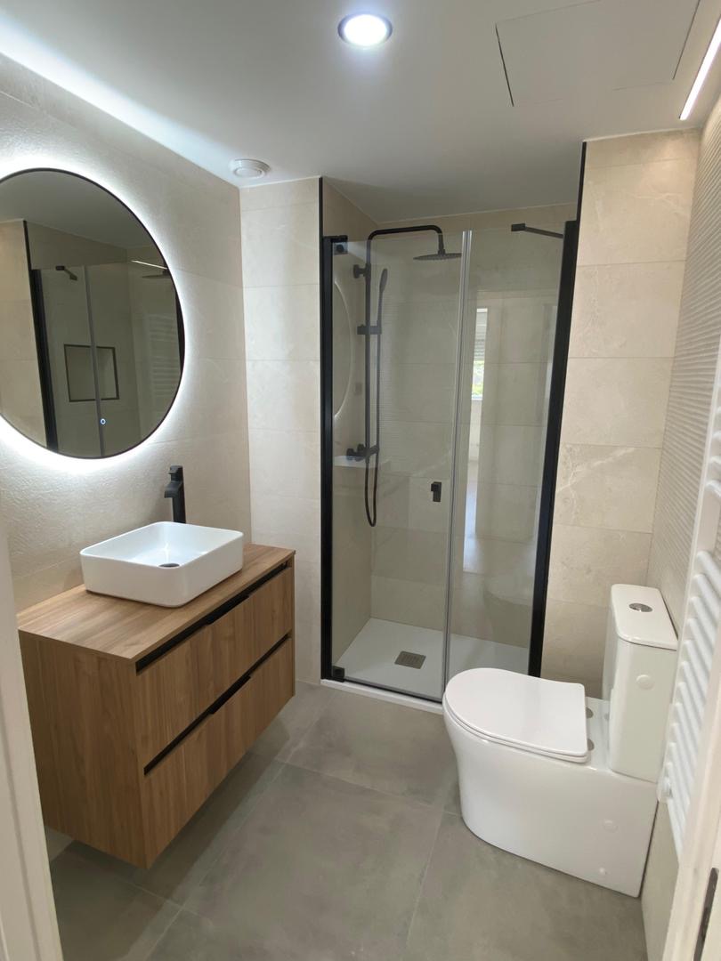 Baño con mampara negra, espejo LED redondo, mueble de madera y lavabo sobre encimera