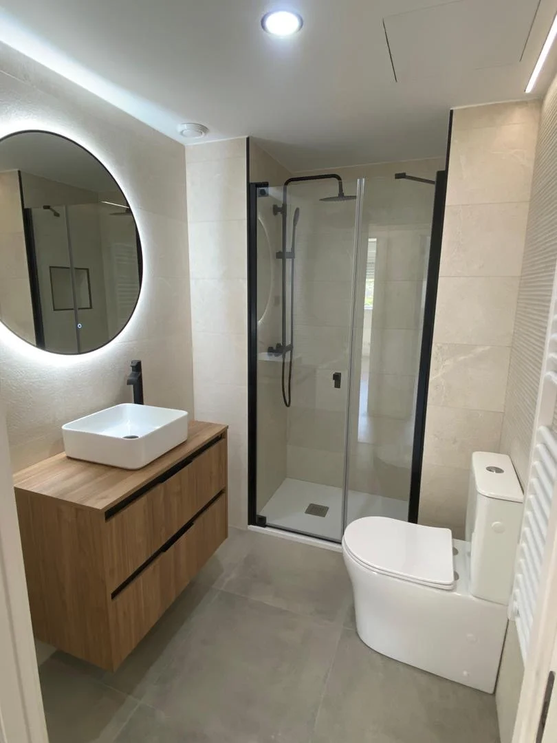 Baño con mampara negra, espejo LED redondo, mueble de madera y lavabo sobre encimera