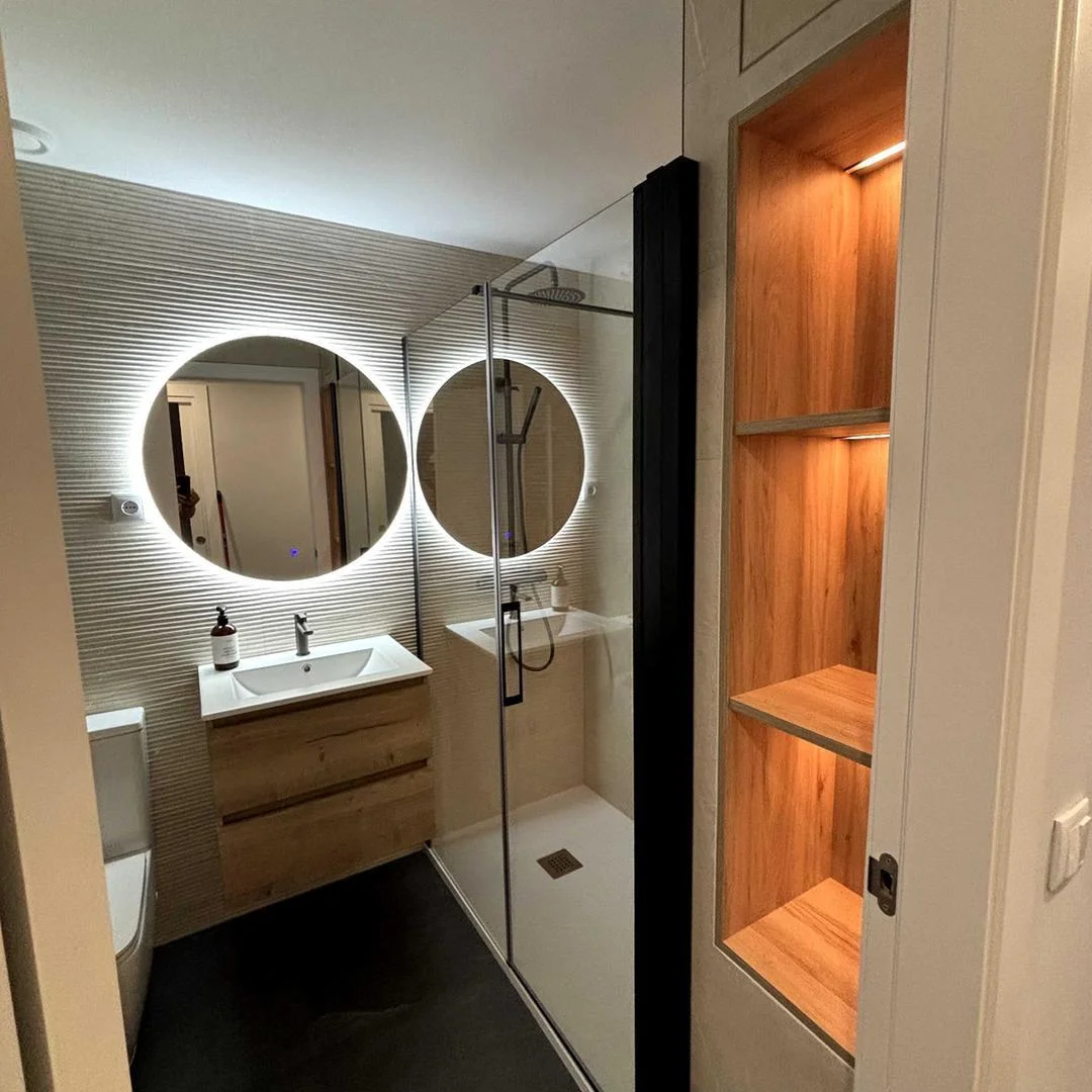 Reforma integral de baño en el Vallès con espejo redondo LED, mueble de madera, ducha de obra y estantería de madera iluminada