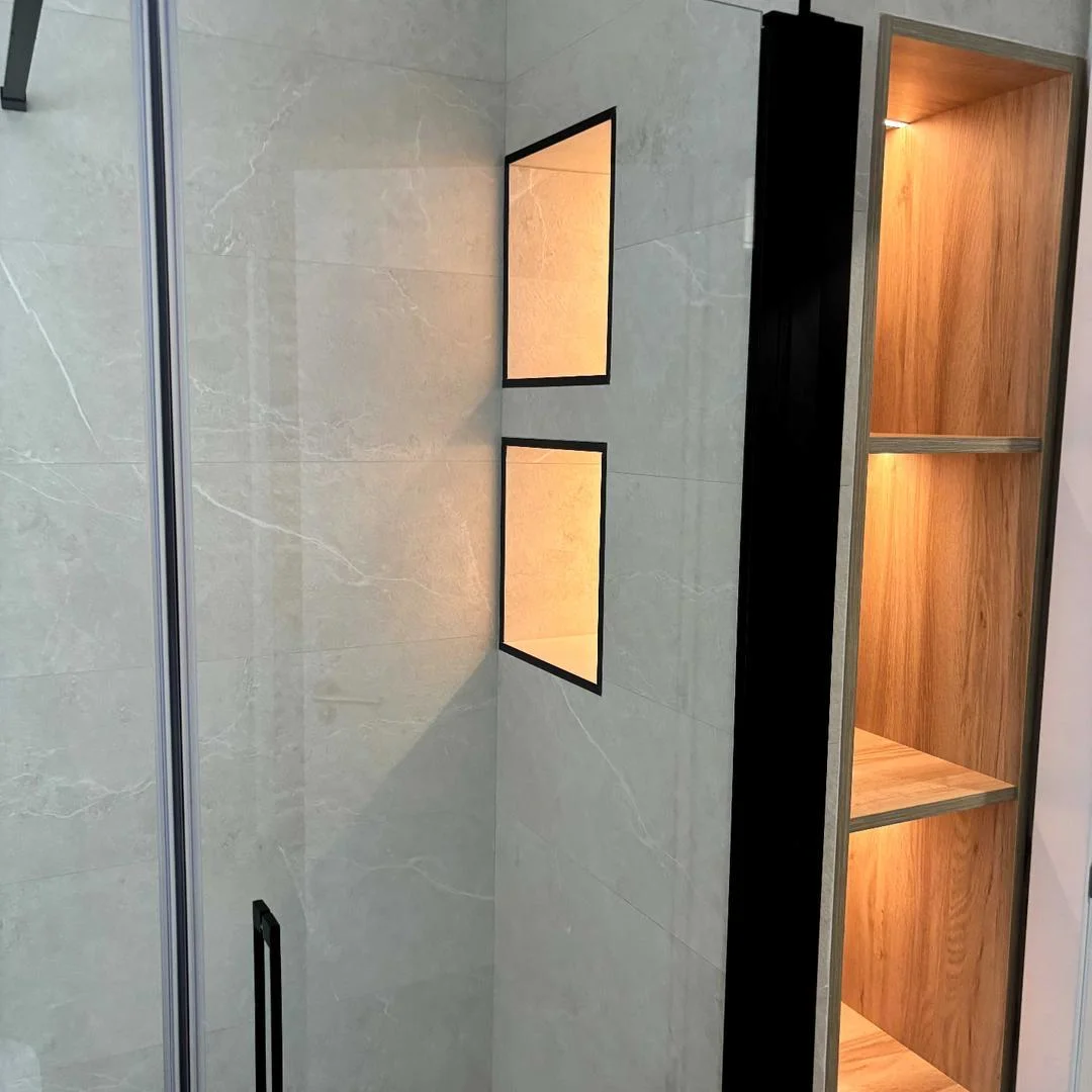 Detalle de ducha con revestimiento de porcelánico gris, hornacinas con marco negro iluminadas y estantería de madera