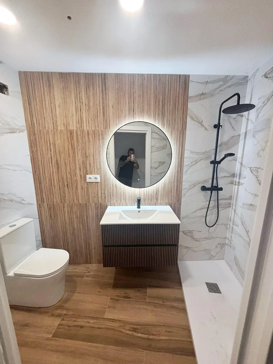 Reforma de baño en el Vallès con pared de lamas de madera, espejo redondo LED, ducha de mármol y grifería negra
