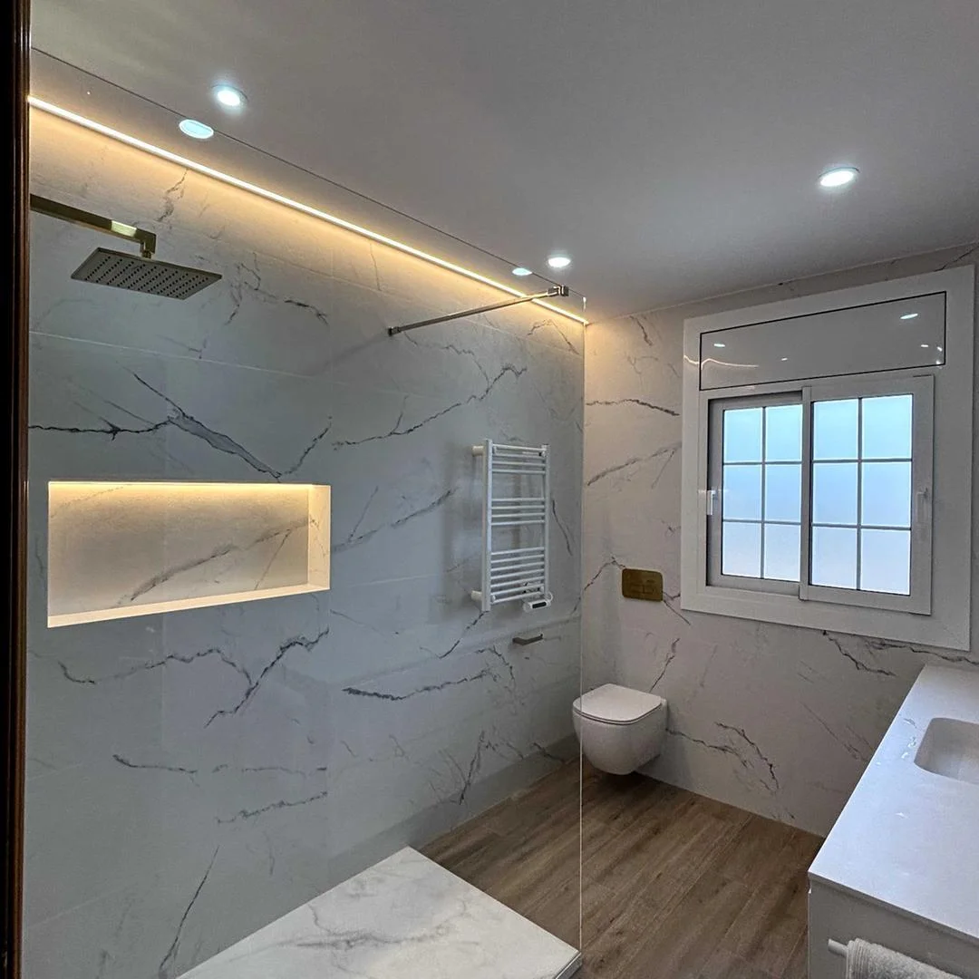 Reforma de baño en Sabadell con revestimiento de mármol blanco, ducha a ras de suelo con grifería dorada e iluminación LED
