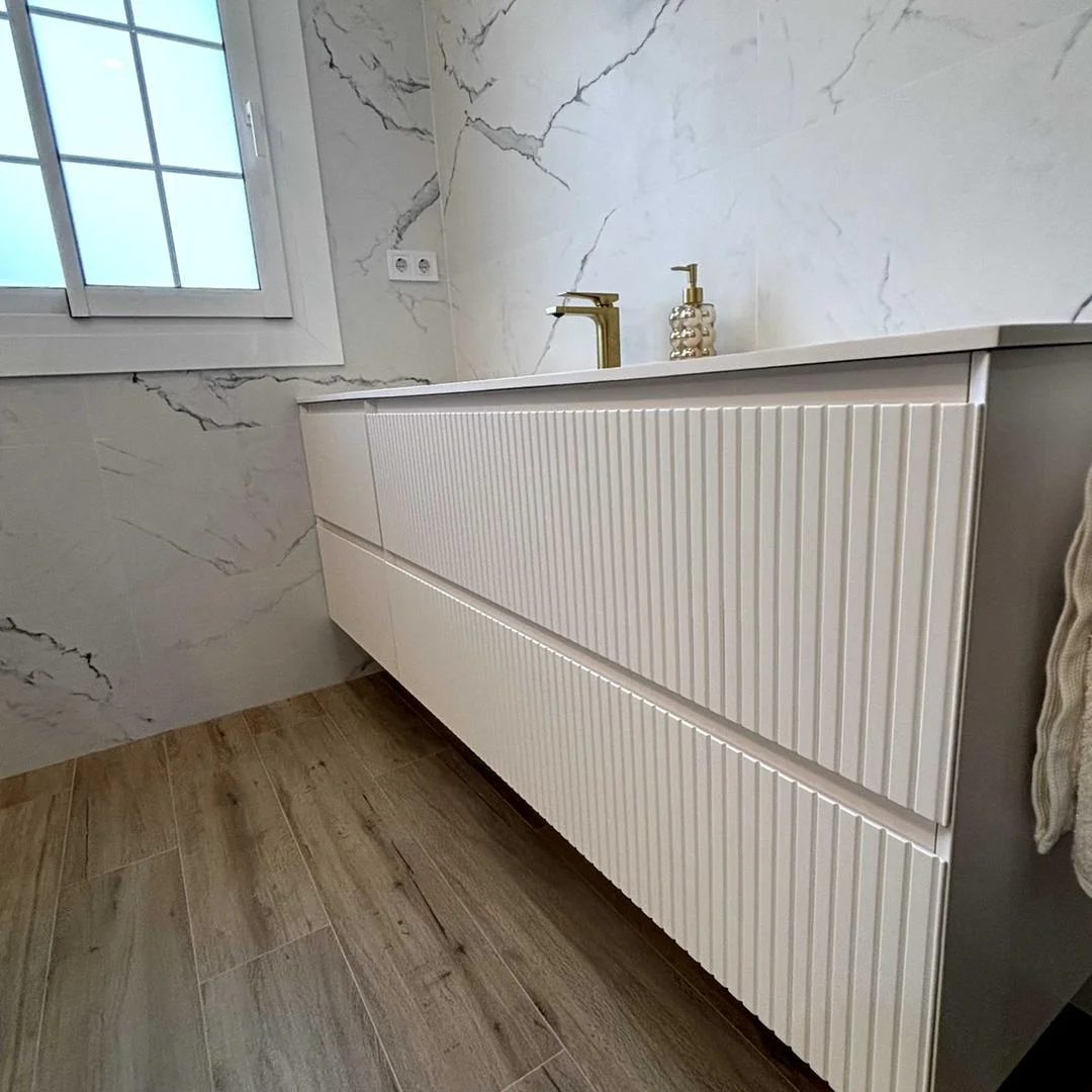 Baño reformado con mueble suspendido de líneas verticales, grifería dorada y revestimiento de mármol blanco