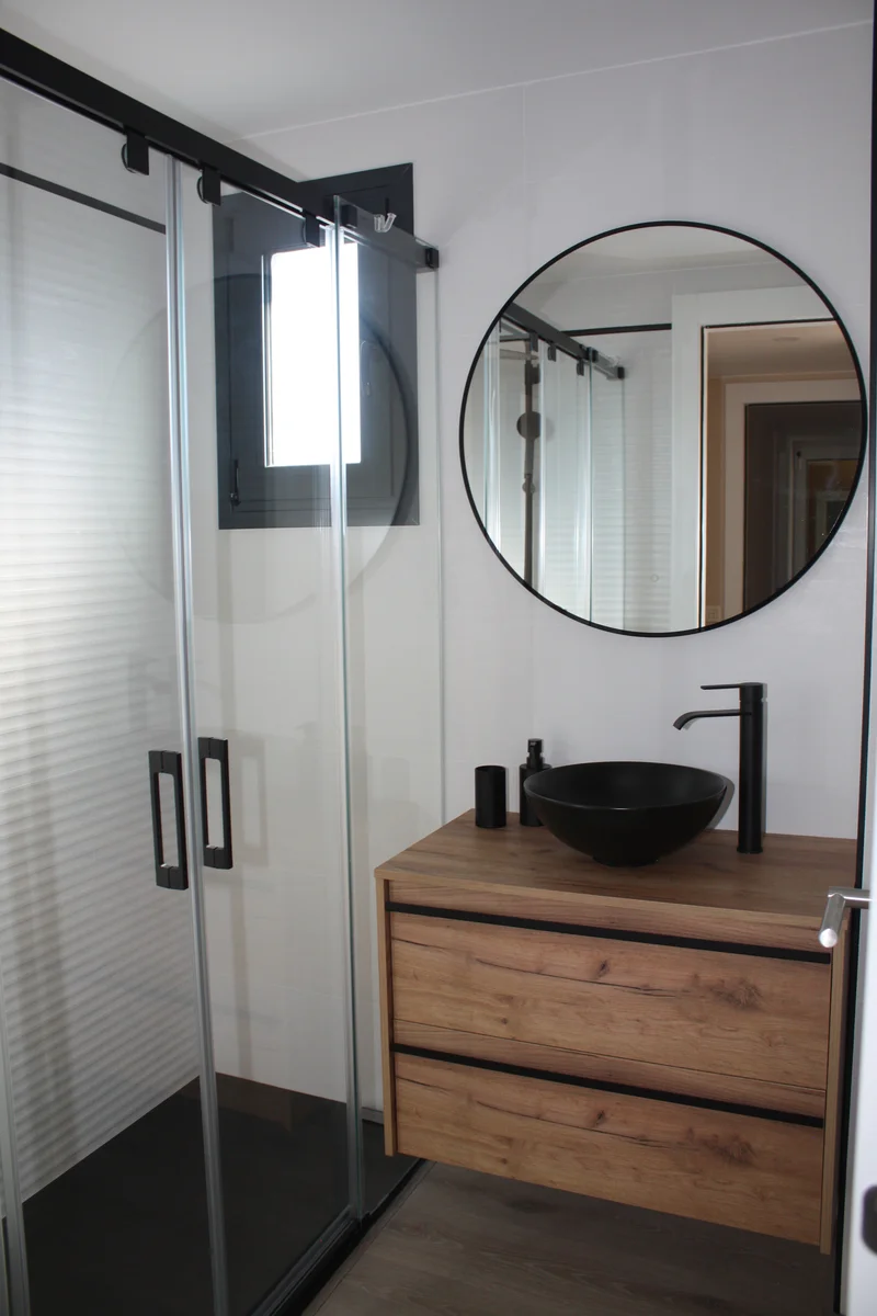 Reforma de baño en Sabadell con ducha de mampara corredera negra, espejo redondo y mueble de madera con lavabo negro